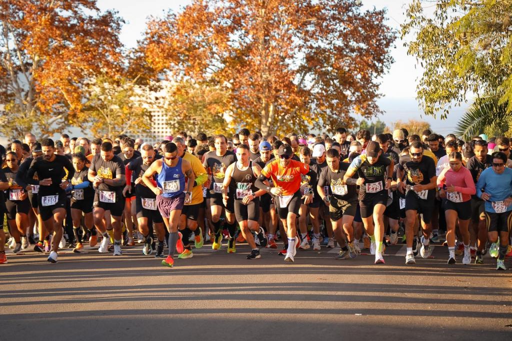 foto colorida horizontal de um grupo de corredores lado a lado na partida da corrida, tendo plátanos iluminados pelo sol ao fundo