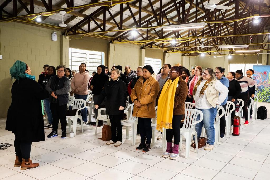 Foto horizontal colorida mostra uma mulher falando para um grupo com cerca de 40 mulheres em um salão comunitário. As participantes estão em pé entre cadeiras brancas, observando a palestrante durante a atividade do curso.