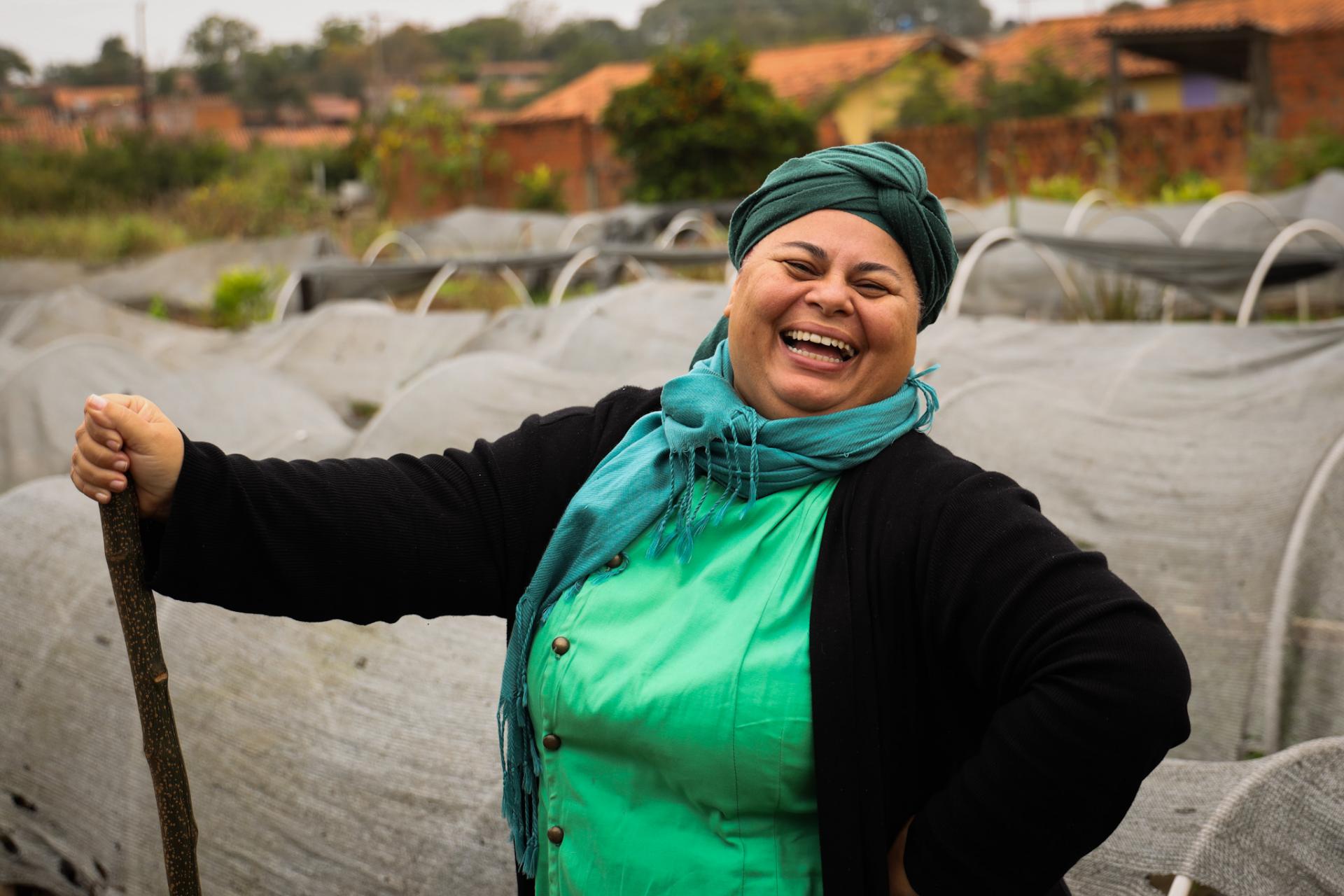 Foto horizontal colorida de uma mulher parda, de estatura média sorrindo ao fundo dá para ver uma horta. Ela usa roupas verdes, cachecol azul-turquesa e turbante escuro, enquanto segura um cabo de ferramenta agrícola.