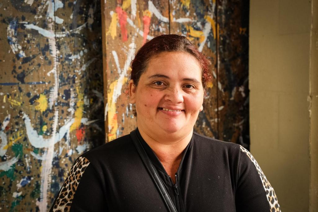 Foto horizontal colorida de uma mulher branca com cabelos vermelhos posando para retrato. Ela sorri para a câmera e veste casaco preto com detalhes estampados nas mangas. Ao fundo, há um painel artístico com manchas coloridas