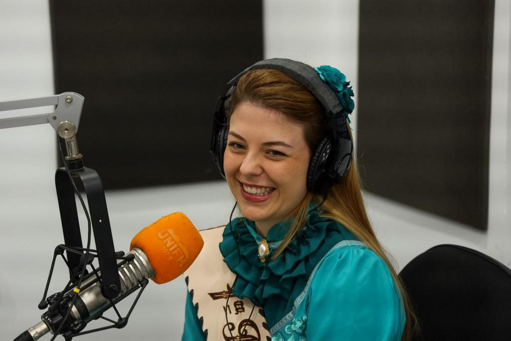 Com fones nos ouvidos, uma menina sorridente fala ao microfone da rádio UniFM. Ela está sentada a uma mesa, tendo como fundo uma parede branca com painéis pretos. Ela é branca e loira, usa um vestido tradicionalista verde e tem pendurada ao ombro a faixa de Primeira Prenda.
