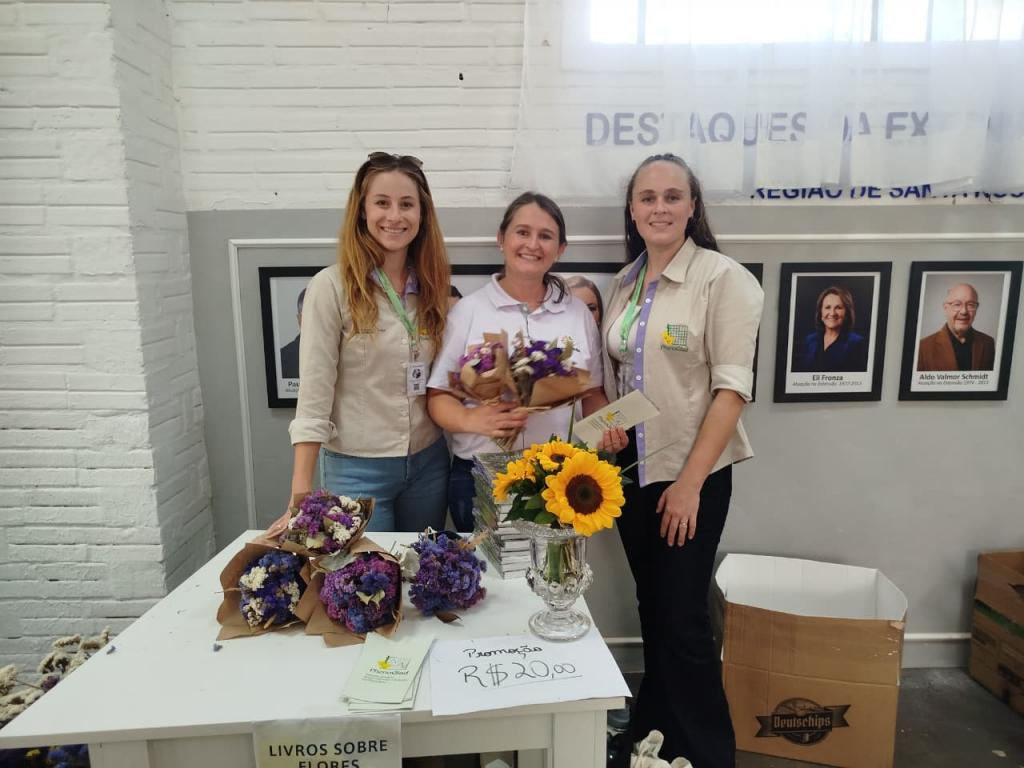 Foto colorida horizontal de três mulheres em estande de feira. Elas seguram girassóis