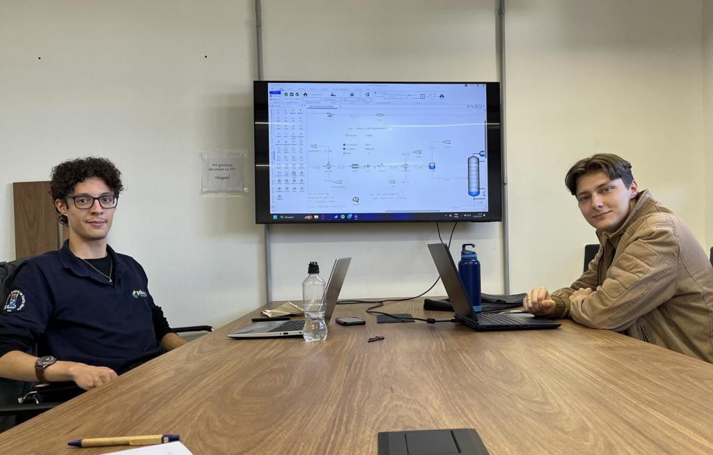 A foto mostra dois rapazes brancos sentados a uma mesa, ambos com computadores à sua frente. O da esquerda usa óculos, tem cabelo curto, crespo e escuro, e veste uma blusa azul-marinho. O da direita veste um casaco bege e tem cabelo curto, liso e loiro. Ao fundo, há uma parede branca com um grande monitor pendurado ao centro, no qual expõem os seus trabalhos.