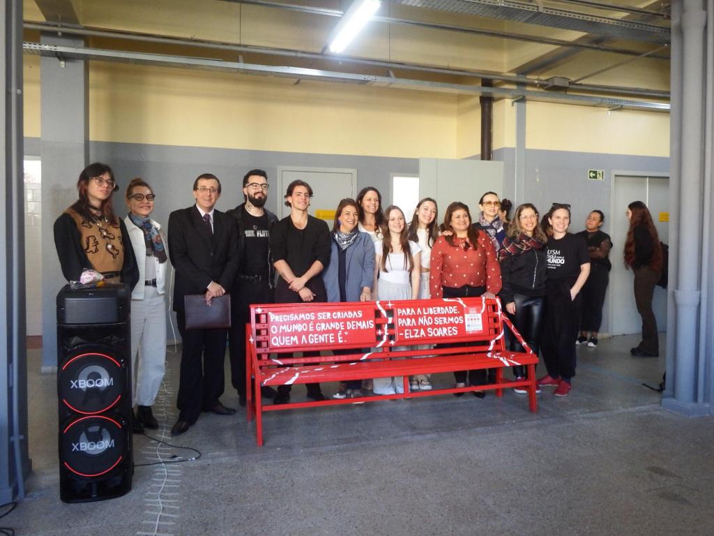 Foto na horizontal, colorida. Em ambiente interno, um grupo de treze pessoas posam para foto atrás do banco pintado de vermelho, colocado ao centro da imagem. No encosto do banco há frases escritas em branco e um cartaz informativo sobre a campanha. As pessoas sorriem e estão lado a lado, algumas com roupas escuras e outras com peças coloridas. À esquerda, há uma caixa de som preta grande no chão. O espaço possui piso cinza, paredes em tons de cinza e bege, colunas laterais e iluminação. Ao fundo, outras pessoas aparecem em pé próximas à parede.