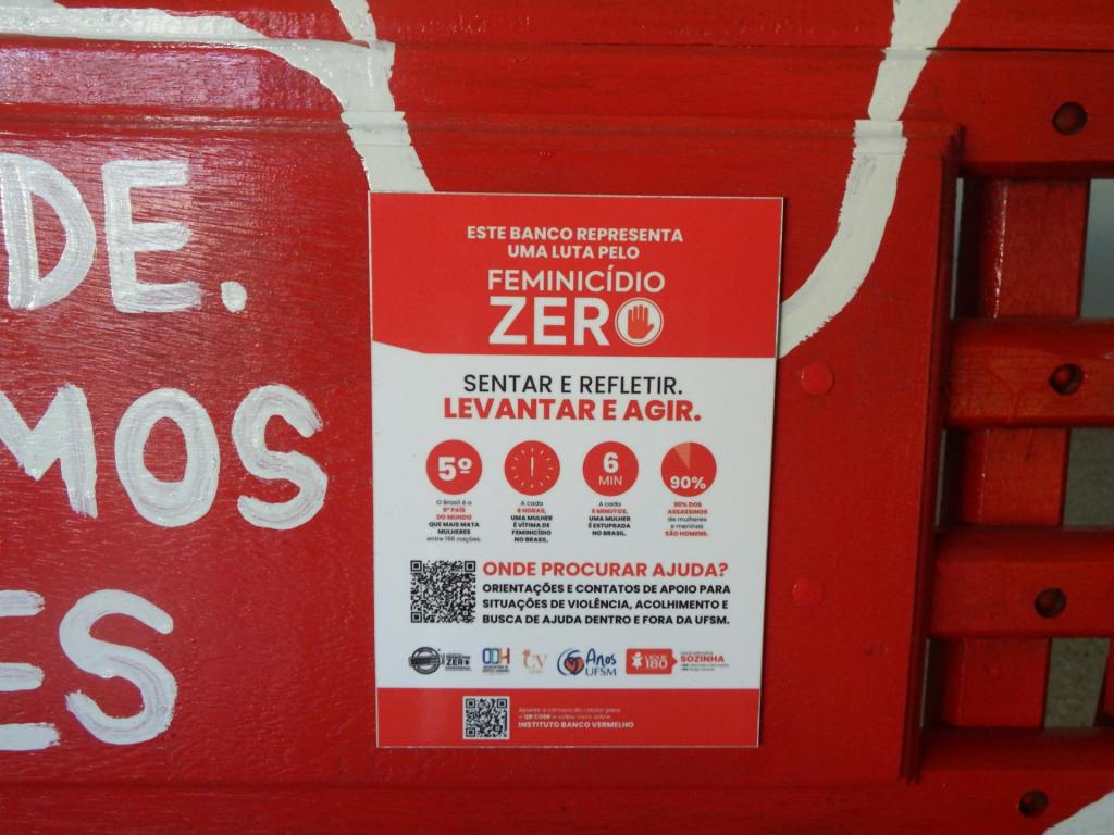 oto colorida na horizontal que mostra, ao centro, um cartaz da campanha “Feminicídio Zero” fixado no banco pintado de vermelho. O cartaz possui fundo branco e vermelho, com textos informativos sobre violência contra a mulher e dados sobre feminicídio no Brasil, além de QR codes e logos institucionais na parte inferior. Ao redor do cartaz, aparecem partes do banco pintadas em tinta branca. À direita, é possível ver a estrutura do encosto do banco em madeira vermelha.