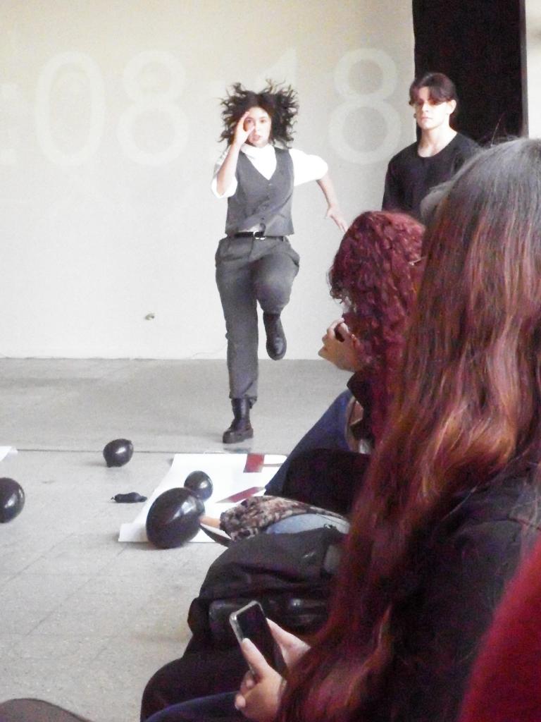 Foto na vertical, colorida. Em ambiente interno, uma estudante se apresenta no centro da imagem durante performance artística. Ela veste roupas cinzas e brancas e está em movimento, com uma perna levantada e um braço próximo ao rosto. Ao fundo, outro estudante de roupa preta acompanha a cena. No chão, há balões pretos e cartazes espalhados. À direita, parte do público aparece sentado, observando a apresentação. O espaço possui piso cinza e parede clara ao fundo.