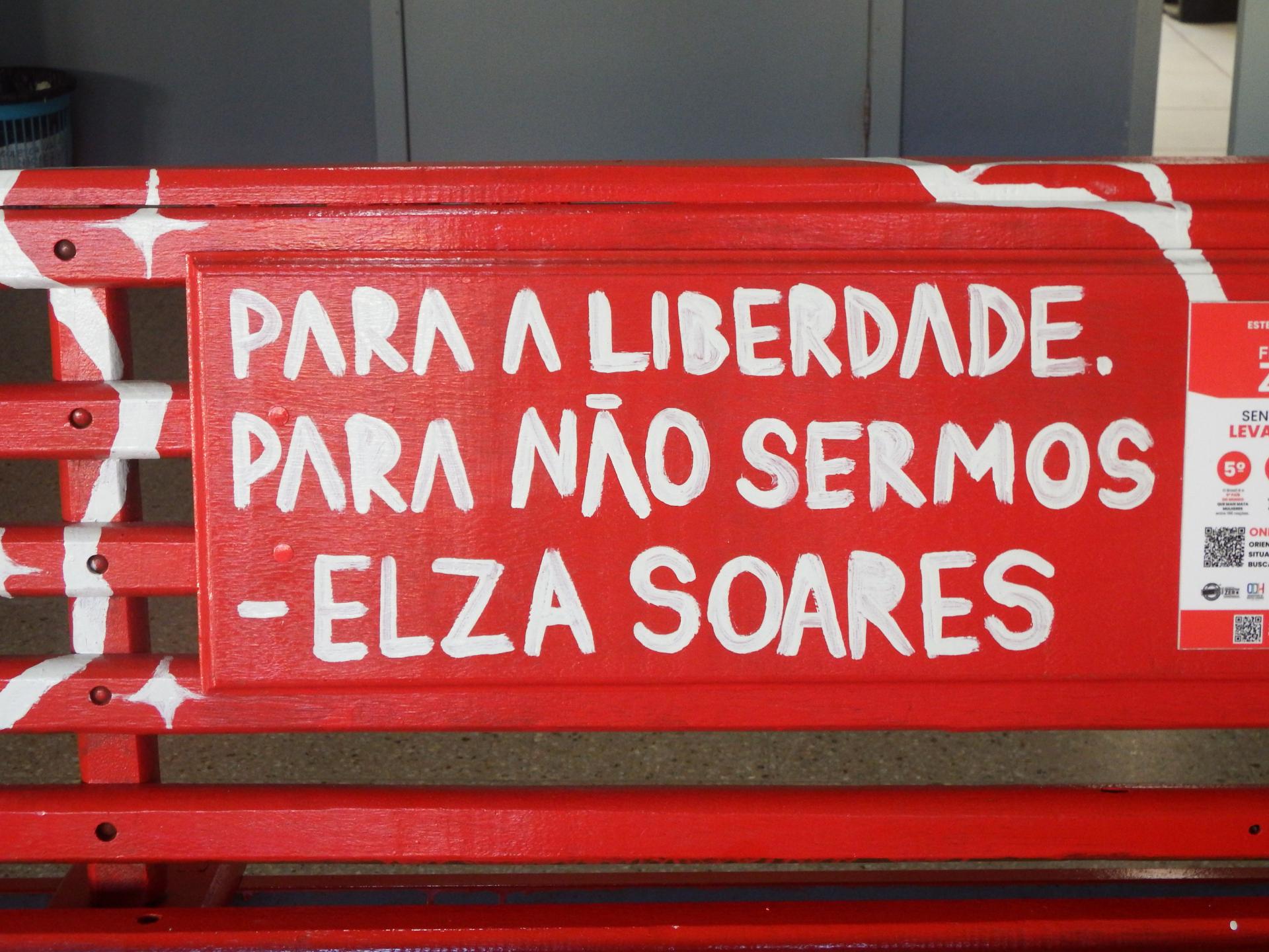 Foto na horizontal, colorida. Em primeiro plano, aparece o banco pintado de vermelho com detalhes em branco. No centro do encosto, há uma placa vermelha com a frase escrita em letras brancas: “Para a liberdade. Para não sermos - Elza Soares”. À direita, parte de um cartaz informativo também vermelho está fixado no banco. Ao fundo, desfocado, há paredes em tons de cinza e parte do piso claro do ambiente interno.