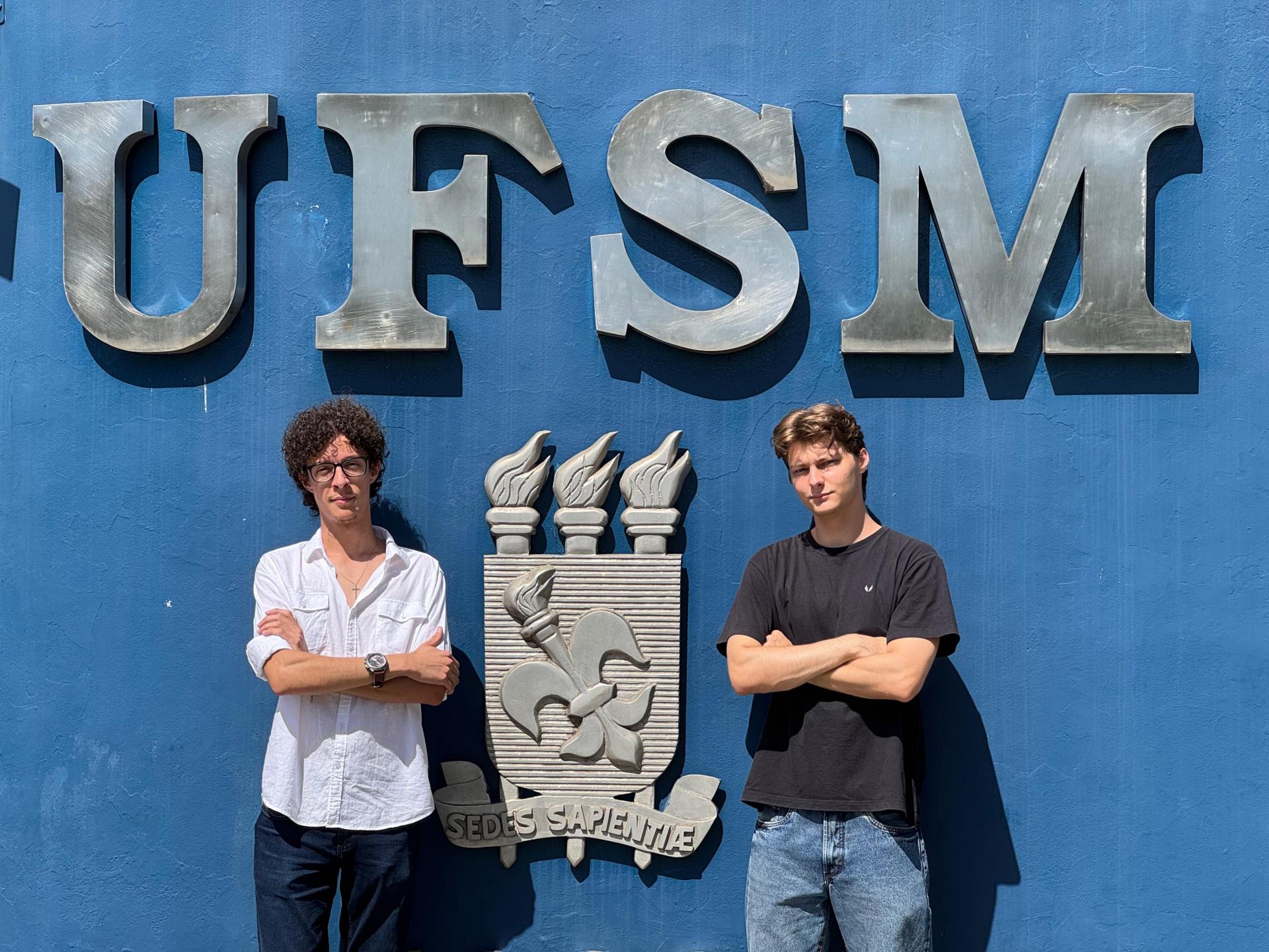 A foto mostra dois rapazes brancos em pé, tendo ao fundo um muro azul com o brasão da UFSM. O da esquerda usa óculos, tem cabelo curto, crespo e escuro, veste calças jeans e uma camisa branca. O da direita tem cabelo curto, liso e loiro, veste calças jeans e uma camiseta preta.