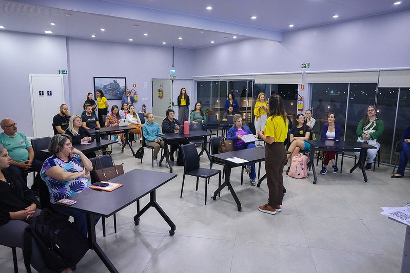 Foto colorida horizontal de várias pessoas sentadas em classes. Em pé, uma mulher com blusa amarela e calça preta