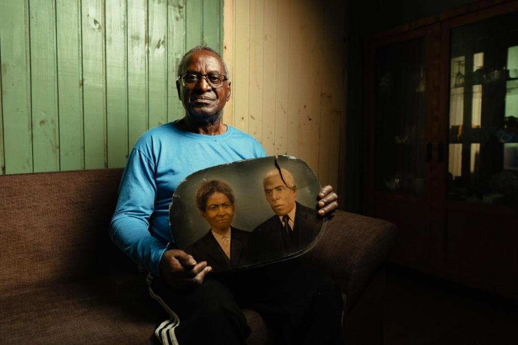 Foto colorida horizontal de homem negro idoso sentado. Ele está segura um retrato de um casal. A fotografia é antiga e tem um formato arredondado.