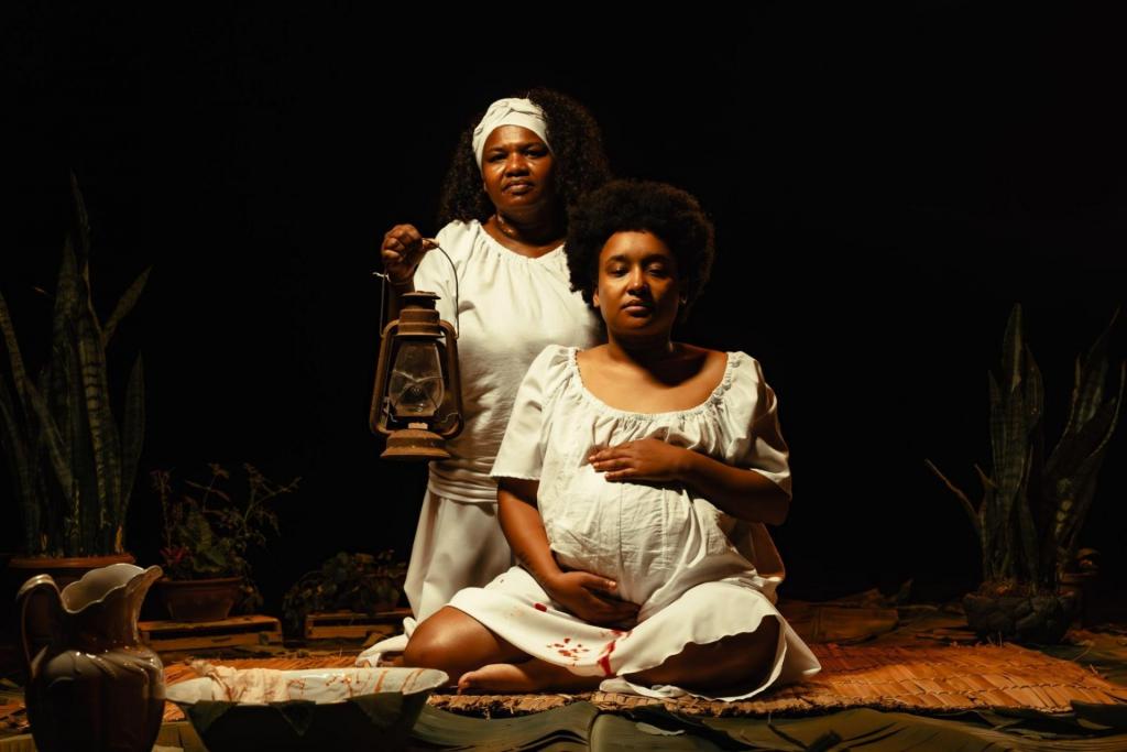 Foto colorida horizal de duas mulheres negras vestidas de branco. A primeira está grávida, sentada e segura a barriga. A segunda está atrás da mulher grávida e segura um lampião. O vestido da mulher grávida apresenta manchas vermelhos.