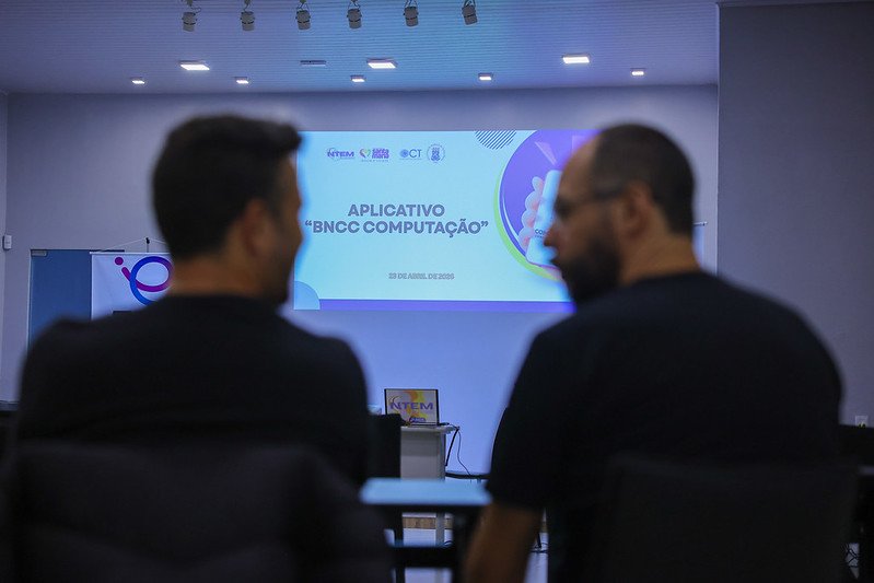 Foto coloorida horizontal de dois homens de costas se olhando. Atrás deles, uma tela com uma projeção sobre o aplicativo