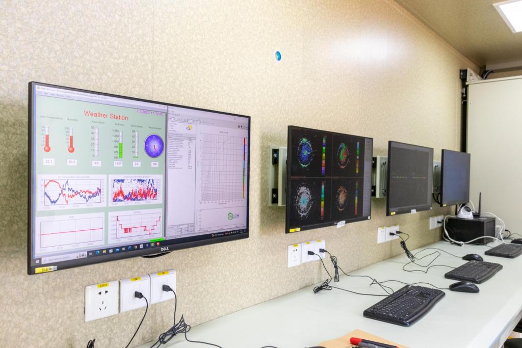 Foto na horizontal colorida mostra uma bancada de laboratório com computadores da área de Meteorologia da UFSM. Ao longo da parede clara, quatro monitores estão alinhados lado a lado acima da mesa. As telas exibem gráficos, mapas, dados meteorológicos e imagens técnicas. No primeiro monitor à esquerda, aparece uma estação meteorológica virtual com indicadores de temperatura, umidade, pressão do ar e velocidade do vento. Sobre a bancada cinza, há teclados, mouses e cabos conectados às tomadas.