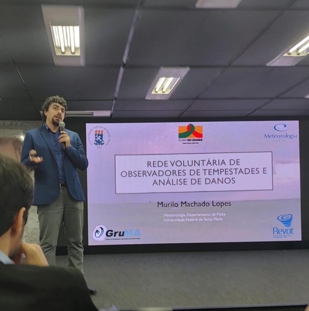 Foto colorida, horizontal. À esquerda, está Murilo Lopes, um homem de cabelos escuros e cacheados, usando blazer azul escuro, camisa escura e calça cinza. Ele aparece em pé sobre o palco segurando um microfone e fazendo uma apresentação. Ao fundo, há um grande painel de projeção. No centro do painel está escrito: “REDE VOLUNTÁRIA DE OBSERVADORES DE TEMPESTADES E ANÁLISE DE DANOS”. Abaixo, aparece o nome “Murilo Machado Lopes” e, em letras menores, “Meteorologia, Departamento de Física, Universidade Federal de Santa Maria”.