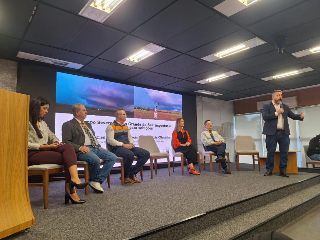 Foto colorida, horizontal. A imagem mostra um palco com cinco participantes sentados em cadeiras. A primeira é uma mulher de cabelos escuros, usando blusa clara, calça vinho e sapatos pretos, olhando para um celular. Ao lado, dois homens sentados usam trajes sociais; um veste paletó escuro com gravata e o outro usa colete em tons escuros e alaranjados sobre camisa clara. Mais à direita, uma mulher de cabelos claros usa roupas em tons escuros com detalhes alaranjados. Ao final da fila, um homem de camisa clara está sentado com as pernas cruzadas, observando a apresentação. À direita, em pé, Vagner Anabor segura um microfone e fala ao público, gesticulando com uma das mãos. Ele veste terno escuro, camisa clara e aparece à frente dos demais participantes. Ao fundo, um grande painel exibe imagens de tempestades e textos relacionados ao evento.