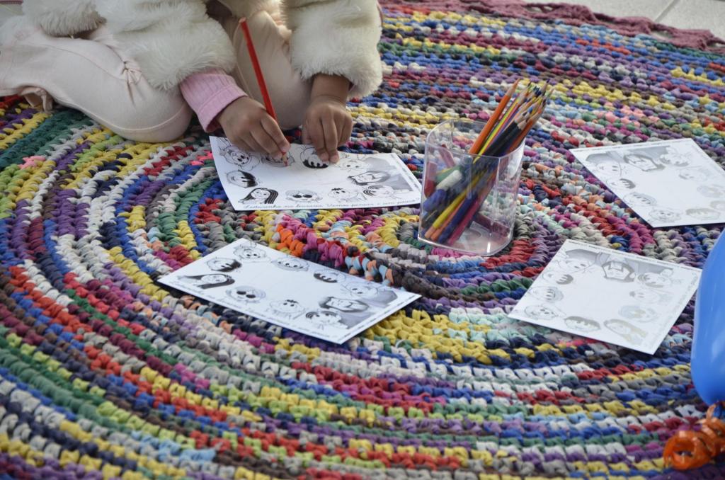 A foto mostra uma criança colorindo desenhos em uma folha. Elá está sentada sobre um tapete colorido estendido no chão, sobre o qual há outras folhas para colorir e vários lápis de cor.