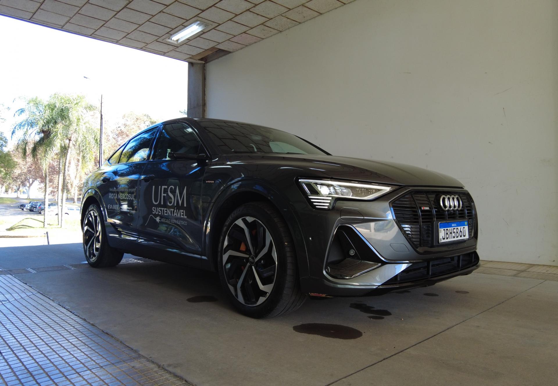 Foto colorida e na horizontal que mostra o carro elétrico da UFSM estacionado em uma área coberta de concreto. O veículo, um Audi cinza-escuro, está posicionado lateralmente, com a parte dianteira voltada para a direita da imagem. Na porta do carro aparecem adesivos com as inscrições “UFSM Sustentável” e referências ao projeto Rota Mercosul de mobilidade elétrica. Ao fundo, à esquerda, é possível ver árvores, gramado e alguns veículos estacionados na área externa iluminada pela luz do dia.