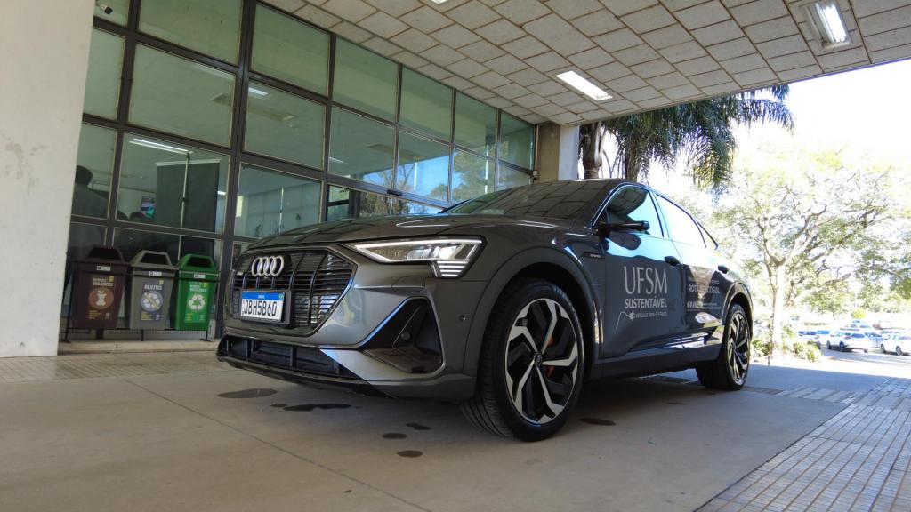 Foto colorida, em formato horizontal. A imagem mostra o veículo elétrico Audi E-Tron estacionado no prédio da Reitoria da UFSM. O carro, na cor cinza-escuro, aparece em destaque em perspectiva frontal lateral, com a identificação “UFSM Sustentável” adesivada na porta. Ao fundo, há uma fachada envidraçada e três lixeiras para coleta seletiva. À direita, árvores e veículos estacionados são vistos na área externa do campus.