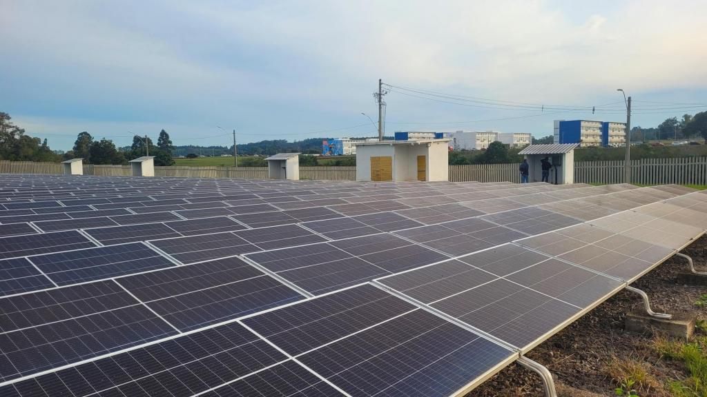 A foto mostra um terreno todo preenchido com painéis solares. Ao fundo, distantes desse local, é possível ver quatro prédios do Campus de Cachoeira do Sul.