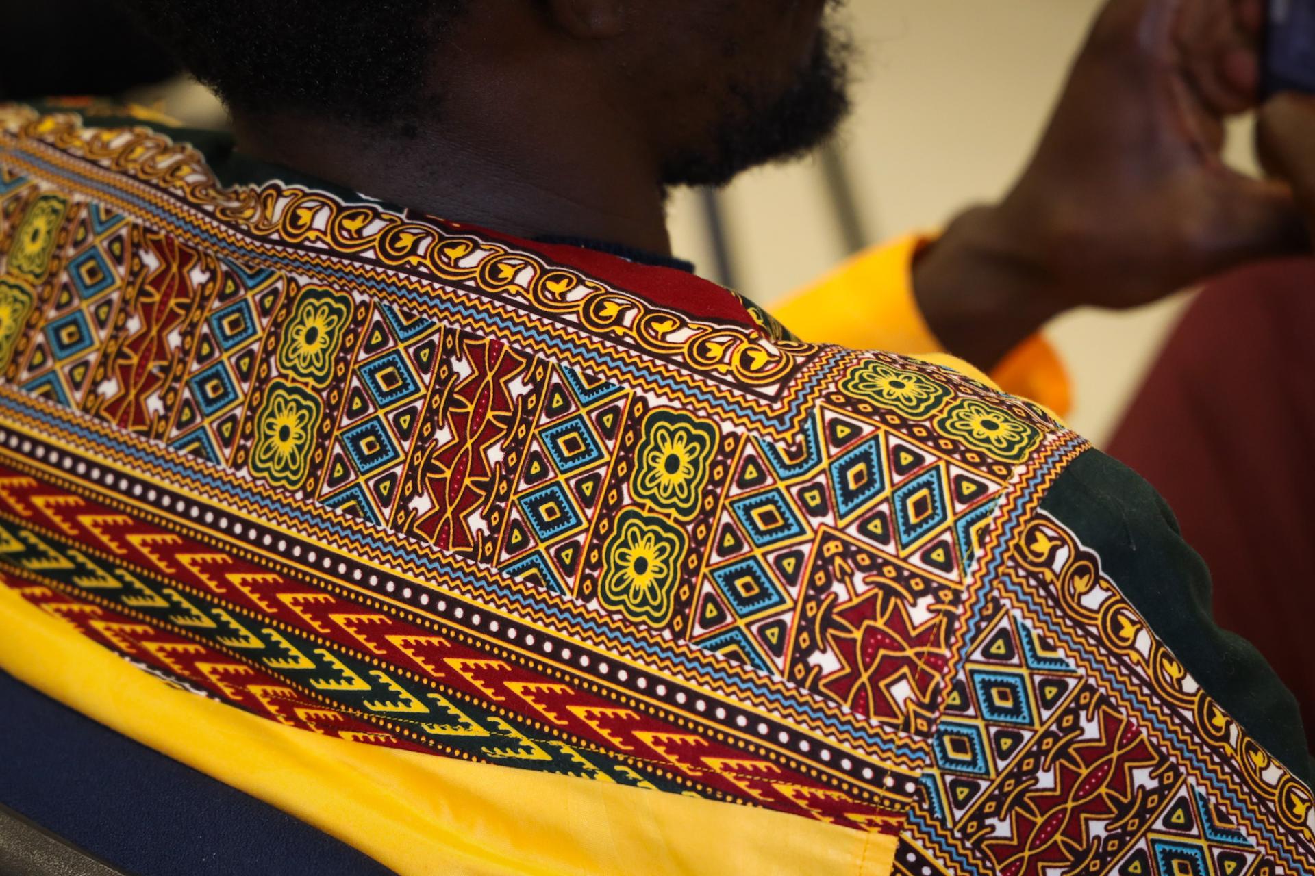 Fotografia em close mostra parte das costas e do ombro de uma pessoa vestindo uma peça de tecido com estampas africanas coloridas. O tecido apresenta padrões geométricos em amarelo, vermelho, azul, preto e branco, com detalhes ornamentais.