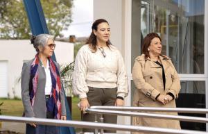 Foto colorida, na horizontal, mostra três mulheres em frente à entrada do novo Restaurante do Colégio Politécnico. Da esquerda para a direita, estão a arquiteta da Pró-Reitoria de Infraestrutura (Proinfra), Gianine Pivetta Mello, a diretora do Colégio Politécnico, Marta Von Ende, e a reitora da UFSM, Martha Bohrer Adaime. As três estão em pé, próximas à porta de vidro do prédio. Em primeiro plano, aparecem os corrimãos metálicos da rampa de acesso ao restaurante. Ao fundo, é possível ver parte da fachada da edificação, com estrutura metálica azul, vegetação e áreas externas do campus.