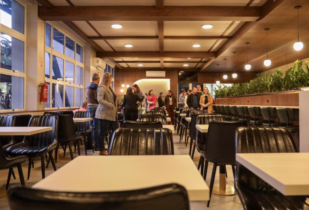 Foto colorida, na horizontal, mostra o interior do novo Restaurante do Colégio Politécnico. O ambiente possui mesas brancas e cadeiras pretas distribuídas ao longo do salão. À esquerda, grandes janelas permitem a entrada de luz natural. O espaço conta com iluminação pendente, revestimentos em madeira e plantas ornamentais que dividem parte da área de atendimento. Ao fundo, um grupo de pessoas circula e conversa durante a visita ao restaurante.