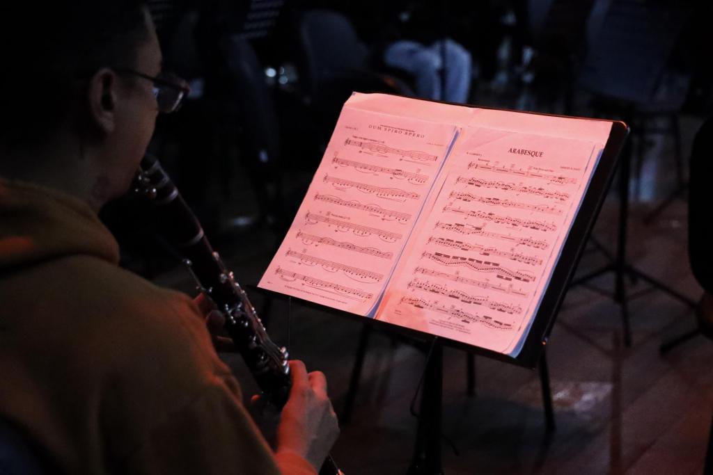 Foto colorida horizontal com pouca luz do palco do Centro de Convenções. A imagem mostra um músico sentado de lado tocando flauta. À frente dele uma partitura em duas páginas