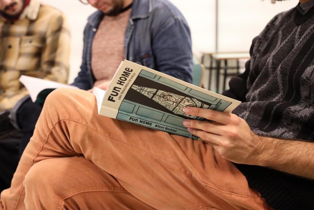 A foto mostra três pessoas sentadas. Em primeiro plano, aparece uma pessoa cujo rosto foi cortado pela foto. Usa calças jeans laranjas e um blusão preto. Está folheando o livro “Fun Home”, de Alison Bechdel. Ao fundo, aparecem dois homens, cujos rostos estão parcialmente cortados.