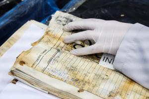 Foto colorida na horizontal mostra uma mão com luva branca, manuseando cuidadosamente um livro de registros danificado pelas enchentes. O documento apresenta páginas amareladas, manchas de umidade, sujeira e sinais de deterioração. Na folha aberta, é possível identificar campos preenchidos à mão e a inscrição “Banco Central do Brasil”. A cena registra uma etapa do trabalho de recuperação e preservação de documentos realizado pelo projeto Hub Doc. Ao fundo, desfocado, aparecem materiais e superfícies utilizados no processo de conservação do acervo.