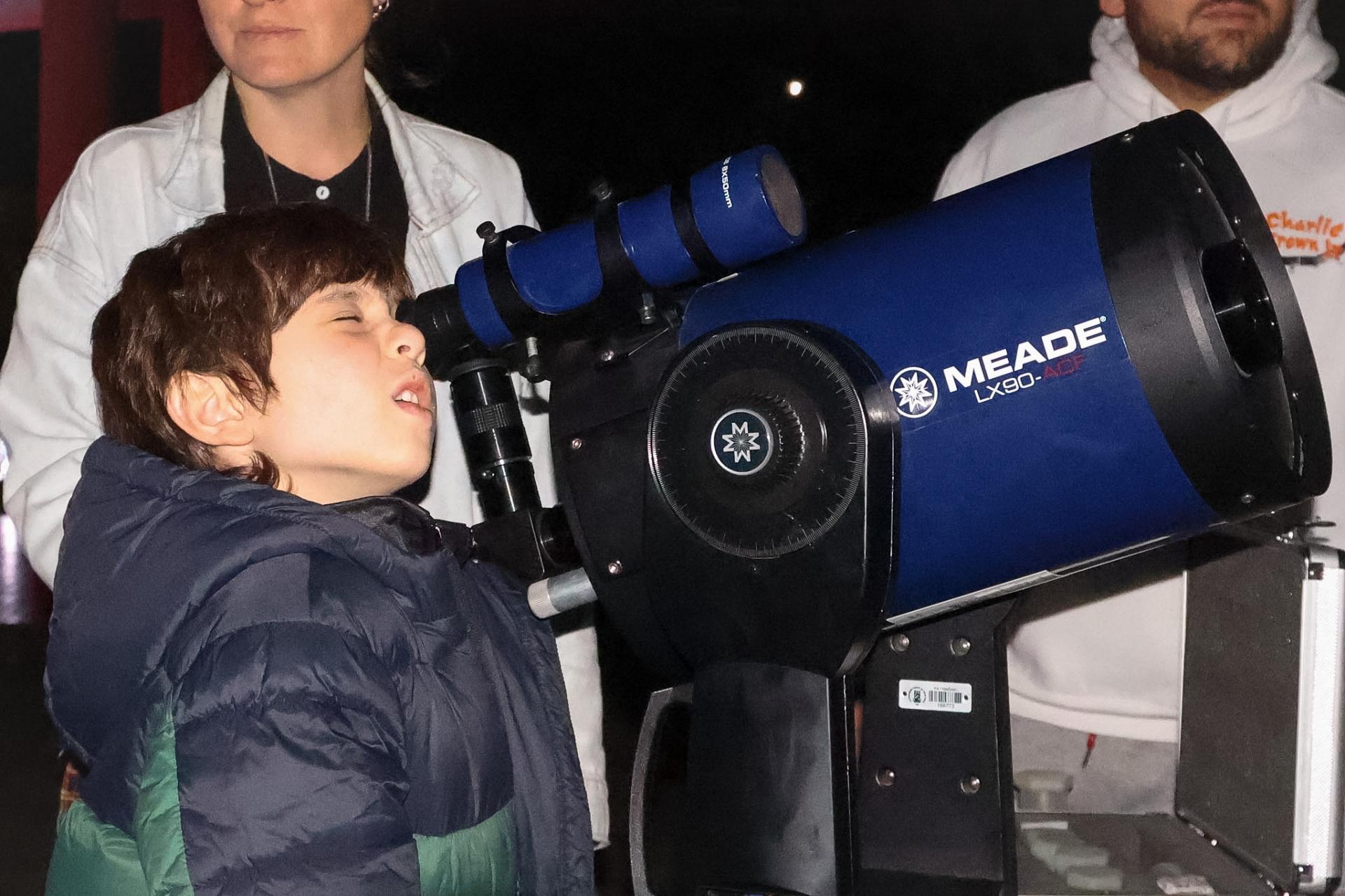 Uma criança observa o céu à noite por meio de um telescópio. É um menino branco, de cabelo castanho, liso e curto. Ele veste um casaco verde e azul. Ao fundo, estão um homem e uma mulher, ambos vestidos de branco. Não é possível ver o rosto inteiro de nenhum dos dois.