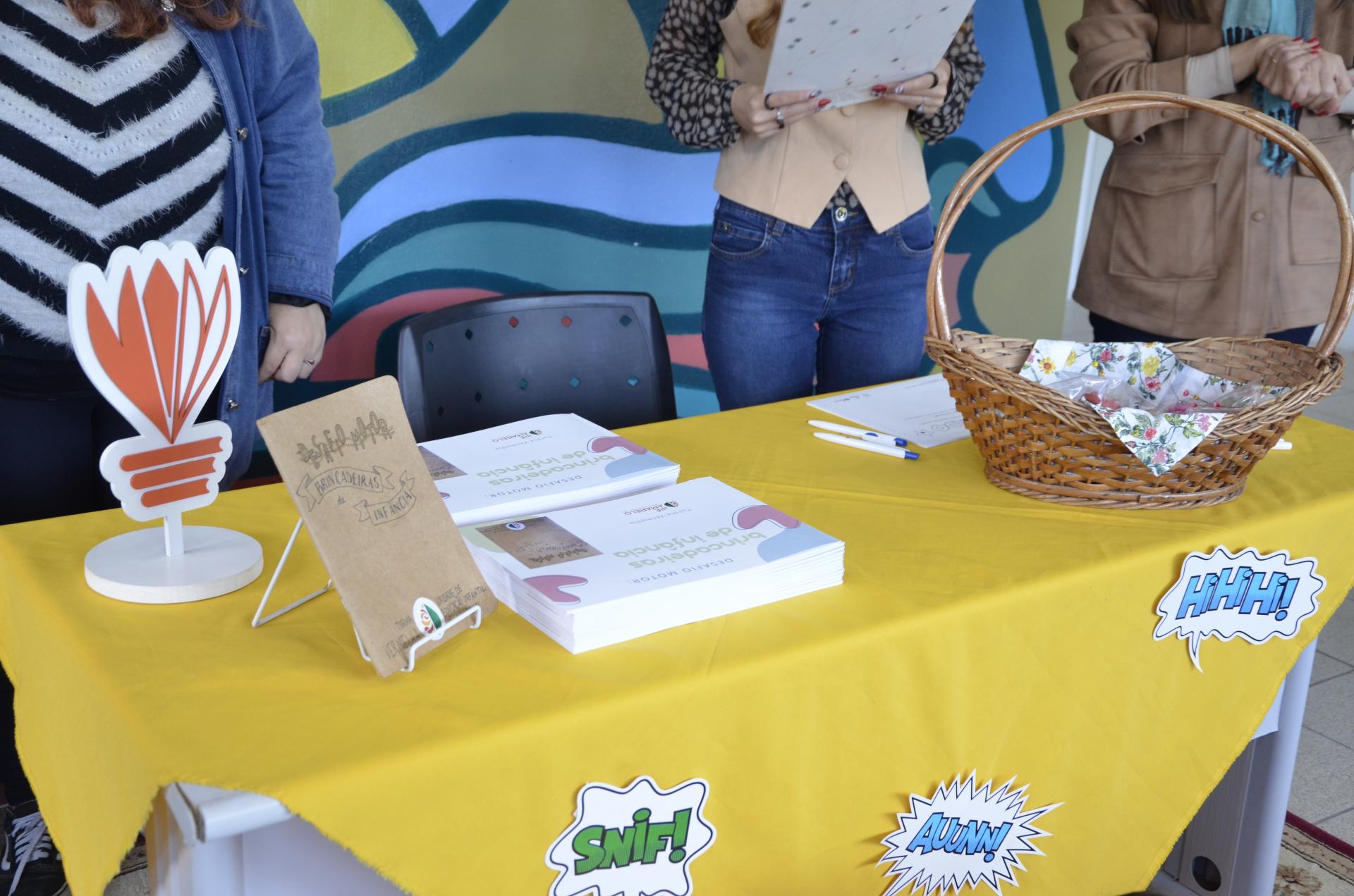 foto colorida horizontal de uma mesa com toalha amarela e livros, uma cesta e outros objetos dispostos sobre ela, com pessoas atrás