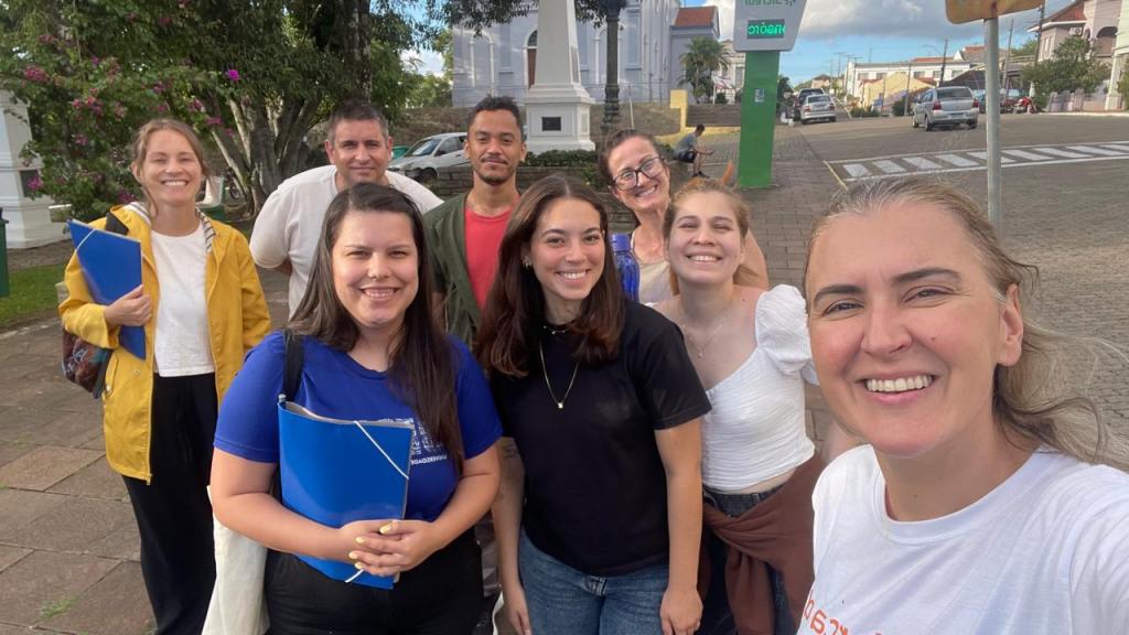 Foto colorida na horizontal. Em primeiro plano, um grupo de oito pessoas sorri para a câmera durante uma atividade de campo do projeto "Comunicação de Proximidade". À direita, a coordenadora Aline registra a imagem em formato de selfie. Ao centro, estudantes e pesquisadores estão reunidos em uma calçada, alguns segurando pastas azuis utilizadas para a aplicação de questionários. Ao fundo, aparecem árvores, uma praça e edificações históricas da região central de Silveira Martins.