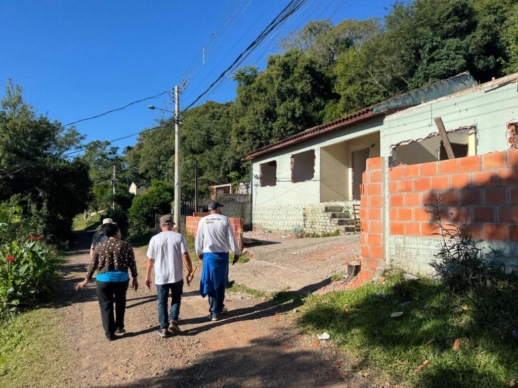Foto colorida na horizontal que exibe uma rua de chão batido, quatro pessoas caminham de costas em direção ao fundo da via. À direita, há uma casa em reforma, com janelas vazadas e com parte das paredes demolidas, além de um muro de tijolos aparentes em construção. Ao redor, árvores densas cobrem uma encosta, enquanto postes e fios elétricos acompanham a rua. O céu está limpo e azul, e a luz do sol ilumina a cena, projetando sombras no chão.