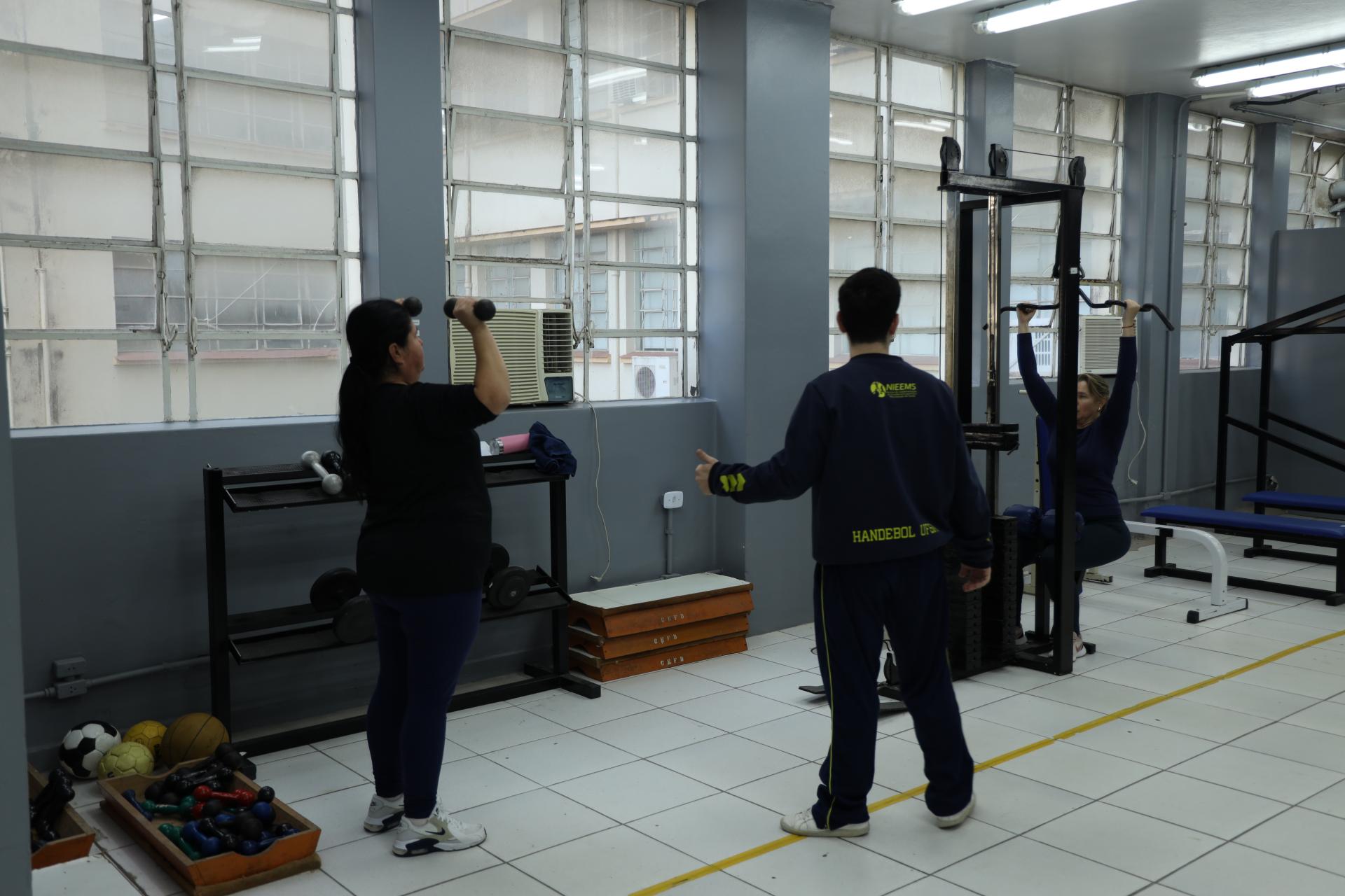 foto colorida horizal da academia popular. À direita, uma mulher em pé segura dois pesos e os levanta para cima. À direita, uma mulher sentada um aparelho de musculação levanta os braços. Ao centro, de costas, o instrutor conversa com ambas