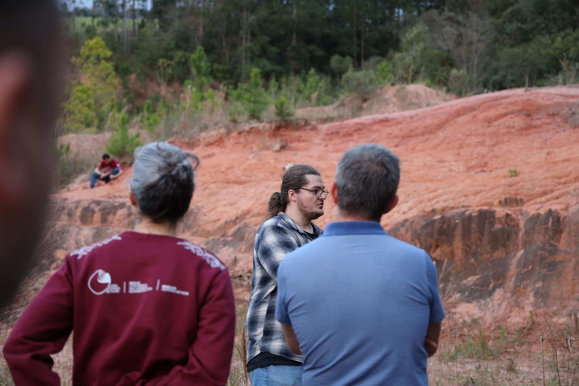 Fotografia colorida na horizontal de uma visita técnica ao Geossítio Fossilífero São Luiz, em Faxinal do Soturno. No centro da imagem, o integrante do CAPPA, Flávio Pretto, está de perfil, usando óculos, camisa xadrez em tons de cinza e preto e calça jeans. Com os cabelos longos presos em um coque, ele conversa com o grupo e apresenta informações sobre o geossítio. Em primeiro plano, de costas para a câmera, está o avaliador do Geoparque Mundial da UNESCO, Jean-Luc, com cabelos curtos grisalhos e camiseta polo azul-clara, acompanhando a explicação. À esquerda, também de costas, aparece uma mulher de cabelos grisalhos presos em um coque, vestindo um moletom bordô com a identidade visual do Geoparque Quarta Colônia. Ao fundo, destaca-se uma extensa formação de solo avermelhado com afloramentos rochosos, característica do geossítio, cercada por vegetação e árvores de médio e grande porte. Sobre a encosta, uma pessoa sentada observa a atividade. A cena acontece ao ar livre, durante o dia, com os participantes voltados para a explicação sobre o patrimônio geológico do local.