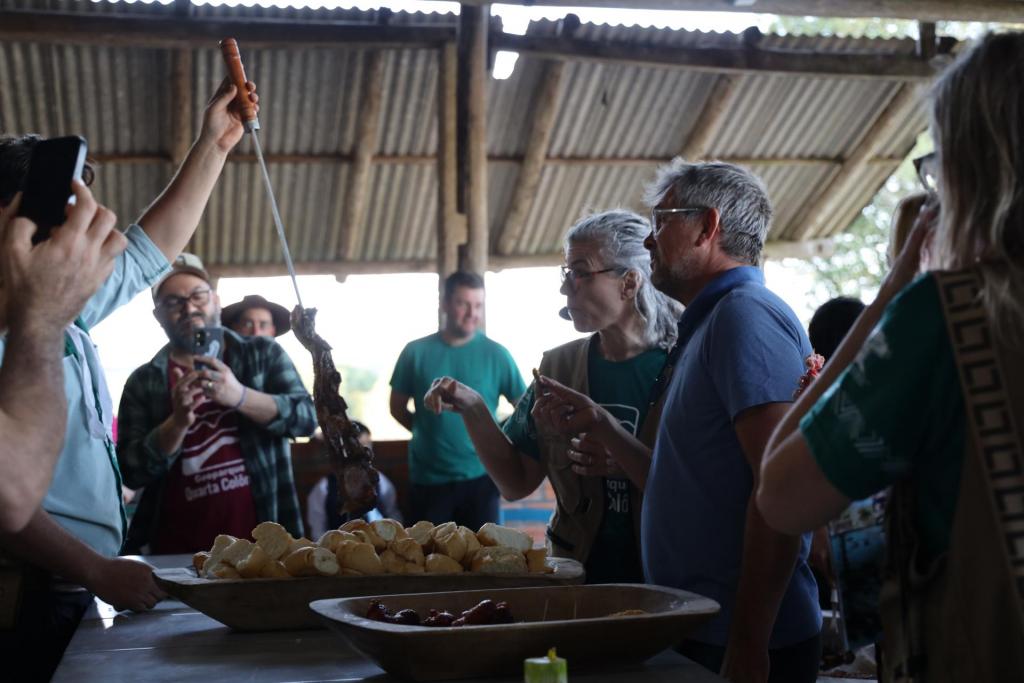 Fotografia colorida na horizontal de um momento de confraternização no CTG Estância do Sobrado, durante a visita dos avaliadores do Geoparque Mundial da UNESCO. No centro da imagem, o avaliador Jean-Luc, um homem de cabelos curtos grisalhos, usa óculos e camiseta polo azul-escura enquanto experimenta um pedaço de churrasco gaúcho. Ao seu lado está a tradutora Amy Lee, uma mulher de cabelos grisalhos presos em um rabo de cavalo, usando óculos, camiseta verde e colete bege. Ela também segura um pedaço de carne e conversa com Jean-Luc. À esquerda, uma pessoa ergue um espeto com um pedaço de carne assada, oferecendo o churrasco aos visitantes. Em primeiro plano, sobre uma mesa, há uma grande travessa de madeira com pães e outra com linguiças, além de outros alimentos típicos. Ao fundo, visitantes acompanham a degustação, alguns registrando o momento com celulares. O ambiente é coberto, com estrutura de madeira e telhado de zinco aparente, característico de um galpão de CTG, iluminado pela luz natural que entra pelas laterais.