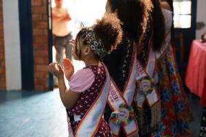 Foto colorida na horizontal. Crianças vestidas com trajes coloridos e faixas atravessadas no peito aguardam em fila próximo à entrada de um salão. A menina em primeiro plano bate palmas enquanto olha em direção ao interior do espaço, iluminado pela luz que entra pela porta.