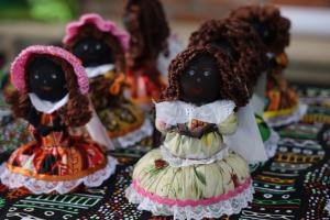 Foto colorida na horizontal. Bonecas artesanais de tecido com diferentes estampas de vestidos e cabelos de lã marrom estão expostas sobre uma mesa coberta por uma toalha com estampa colorida de inspiração africana. A imagem destaca uma boneca em primeiro plano, enquanto as demais aparecem desfocadas ao fundo