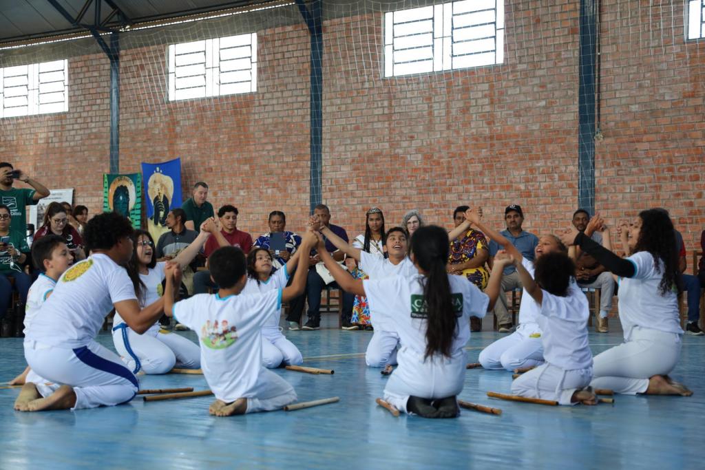 Fotografia colorida na horizontal de uma apresentação cultural realizada por crianças do Quilombo Vovó Isabel para os avaliadores do Geoparque Mundial da UNESCO e o público. No centro da imagem, cerca de dez crianças vestidas com camisetas e calças brancas formam um círculo sobre o piso azul de um ginásio. Ajoelhadas no chão, elas mantêm os braços erguidos e as mãos dadas, enquanto algumas cantam ou olham para o alto, durante a apresentação. No chão, ao redor do grupo, estão distribuídos bastões de madeira utilizados na atividade. Ao fundo, uma fileira de cadeiras reúne os avaliadores e demais participantes, que acompanham atentamente a apresentação. Entre eles, a coordenadora do Quilombo Vovó Isabel, Joceli Pereira, com vestido amarelo estampado, além de outros moradores e visitantes. Algumas pessoas registram o momento com celulares. O espaço possui paredes de tijolos aparentes, janelas altas com entrada de luz natural e cobertura metálica. Atrás do público, dois painéis coloridos com referências à cultura afro-brasileira complementam o ambiente.
