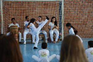 Foto colorida na horizontal. Criança realiza um movimento de capoeira no centro de uma roda formada por músicos e participantes vestidos de branco, em um ginásio. Ao fundo, há uma trave de futebol com rede e uma parede de tijolos aparentes. Os músicos tocam atabaque, berimbau e pandeiro, enquanto outras crianças assistem sentadas no chão em primeiro plano