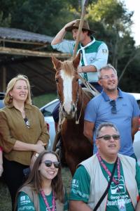 Foto colorida na vertical. Homem montado em um cavalo segura um bastão e ajusta o chapéu durante uma atividade ao ar livre. Em frente ao cavalo, um homem e uma mulher observam a cena sorrindo. Ao fundo, aparecem árvores, um galpão rural e veículos estacionados.
