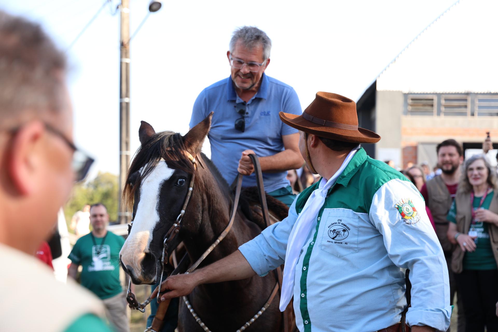 Fotografia colorida na horizontal da recepção ao avaliador do Geoparque Mundial da UNESCO, Jean-Luc, no CTG Estância do Sobrado. No centro da imagem, Jean-Luc está montado em um cavalo de pelagem escura com uma larga faixa branca no rosto. Ele tem cabelos curtos grisalhos, usa óculos e veste camiseta polo azul-clara, enquanto sorri e olha para o homem que segura as rédeas do animal. À frente do cavalo, de costas para a câmera, está um homem com chapéu marrom de aba larga, lenço branco no pescoço e camisa azul-clara com detalhes verdes. Na manga esquerda da camisa há um brasão bordado, e no bolso frontal está bordado o nome CTG Estância do Sobrado – Pinhal Grande (RS). Com a mão direita, ele segura as rédeas do cavalo. Ao redor da cena, diversas pessoas acompanham a recepção. No primeiro plano, à esquerda, aparecem desfocadas as cabeças e os ombros de duas pessoas. Ao fundo, há visitantes observando o momento, alguns com camisetas verdes e outros usando crachás. Também são visíveis parte de um galpão com telhado metálico, um poste de iluminação e o céu claro