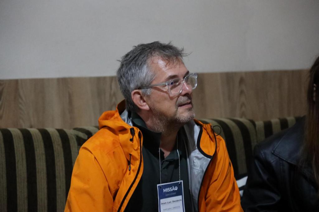 Fotografia colorida na horizontal do avaliador da Unesco Jean-Luc Desbois durante entrevista no Sítio Chaleira Preta, em Caçapava do Sul. No centro da imagem, Jean-Luc está sentado em um sofá listrado, com o corpo voltado para a direita e o olhar direcionado a outra pessoa. Ele tem cabelos grisalhos curtos, barba, usa óculos de armação transparente, jaqueta impermeável laranja sobre uma camisa verde e um crachá de identificação pendurado no pescoço. Ao fundo, há uma parede em tons de branco e madeira. À direita da imagem, aparece parcialmente outra pessoa sentada ao seu lado.