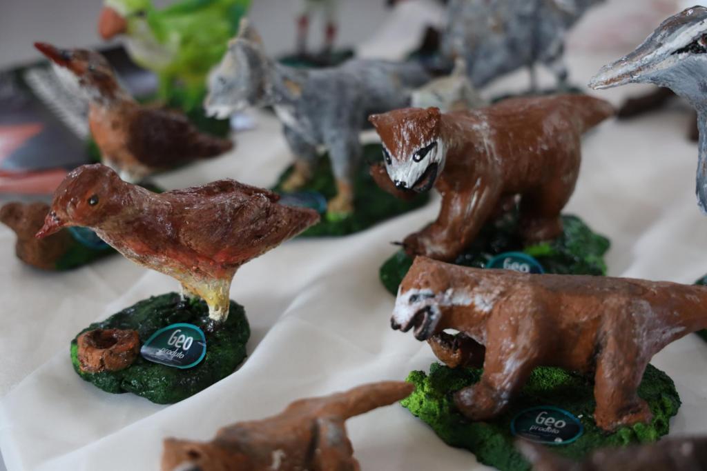 Fotografia colorida na horizontal de geoprodutos artesanais expostos na Feira Municipal da Agricultura Familiar e Produtos Coloniais (Femapro), em Caçapava do Sul. Em primeiro plano, sobre uma mesa coberta por um tecido branco, estão miniaturas de animais da fauna local produzidas em papel machê. À esquerda, destaca-se a figura de um pássaro marrom com detalhes em tons avermelhados, apoiado sobre uma base verde que exibe um selo com a inscrição "Geoproduto". À direita, aparecem duas esculturas de mamíferos marrons, também sobre bases verdes com o mesmo selo. Ao centro da imagem, em segundo plano, há outras miniaturas, entre elas um lobo-guará em tons de cinza e um pássaro de plumagem verde. À direita, aparece parcialmente a escultura de uma ave cinza com bico comprido. As peças representam animais encontrados na região de Caçapava do Sul e fazem parte dos geoprodutos comercializados na feira.
