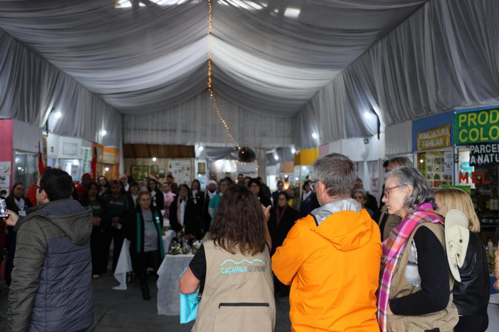 Fotografia colorida na horizontal da visita dos avaliadores da Unesco à Feira Municipal da Agricultura Familiar e Produtos Coloniais (Femapro), em Caçapava do Sul. À esquerda da imagem, um homem de costas, com casaco cinza e verde, registra o momento com um telefone celular. No centro, uma integrante da equipe do Geoparque Caçapava, de costas, veste um colete bege com a inscrição "Caçapava Geoparque" e aponta em direção ao grupo reunido. Ao lado dela, o avaliador Jean-Luc também está de costas, veste uma jaqueta impermeável laranja e acompanha a apresentação. À direita, Amy Lee, tradutora da missão, tem cabelos grisalhos, usa óculos, colete bege e cachecol xadrez em tons de rosa e observa atentamente a atividade. Ao lado dela, Daniela Rocha aparece parcialmente visível, com cabelos loiros e jaqueta preta. Ao fundo, dezenas de pessoas formam uma roda em torno de mesas de exposição enquanto acompanham a fala da equipe do Geoparque. O ambiente é um pavilhão coberto, com teto revestido por tecido branco e uma fileira de pequenas luzes no centro da cobertura. Nas laterais, encontram-se os estandes da feira. À direita da imagem, aparecem placas com as inscrições "Agricultura Familiar", "Produtos Coloniais", "Parceria com a Natureza" e "Mel", além de prateleiras com produtos coloniais expostos. A iluminação combina luz natural e luminárias distribuídas ao longo do pavilhão.