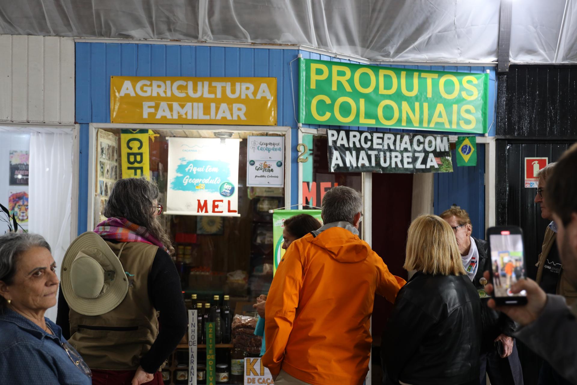 Fotografia colorida na horizontal da visita dos avaliadores da Unesco à Feira Municipal da Agricultura Familiar e Produtos Coloniais (Femapro), em Caçapava do Sul. No centro da imagem, o avaliador Jean-Luc, de costas, veste uma jaqueta impermeável laranja e observa os geoprodutos expostos em um estande. À sua direita, Daniela Rocha, com cabelos loiros na altura dos ombros e jaqueta preta, também está de costas e acompanha a visita. Em frente aos dois, feirantes conversam com o grupo, parcialmente encobertos pelos visitantes. À esquerda da fotografia, uma mulher de cabelos grisalhos, blusa azul e brincos dourados olha em direção à câmera. À frente dela, Amy Lee, a tradutora, aparece de costas, usa um colete bege, cachecol em tons de rosa e chapéu de aba larga preso às costas. Sobre uma pequena bancada, há garrafas de bebidas artesanais, potes e outros produtos coloniais. Ao fundo, o estande é pintado em azul e exibe placas escritas "Agricultura Familiar" e "Produtos Coloniais", além de uma faixa com a frase "Parceria com a Natureza". Na vitrine há cartazes sobre geoprodutos e produtos coloniais, incluindo uma placa com a inscrição "Aqui tem Geoproduto" e outra indicando a venda de mel. À direita da imagem, uma pessoa registra a visita com um telefone celular que mostra o grupo reunido em frente ao estande. O ambiente é coberto e iluminado por luz artificial, com outros visitantes circulando ao redor da feira.