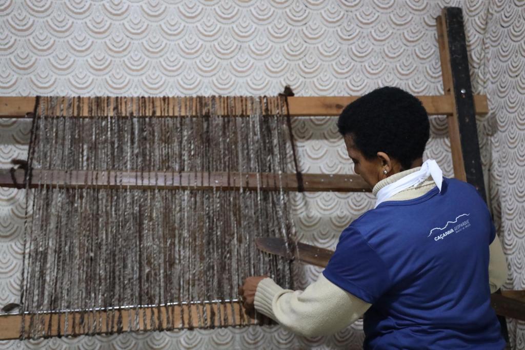 Foto colorida horizontal de mulher de costas trabalhando em um tear de lã. Ela mostra como é tramada a lã de ovelha para produzir uma peça usada na sela de cavalo