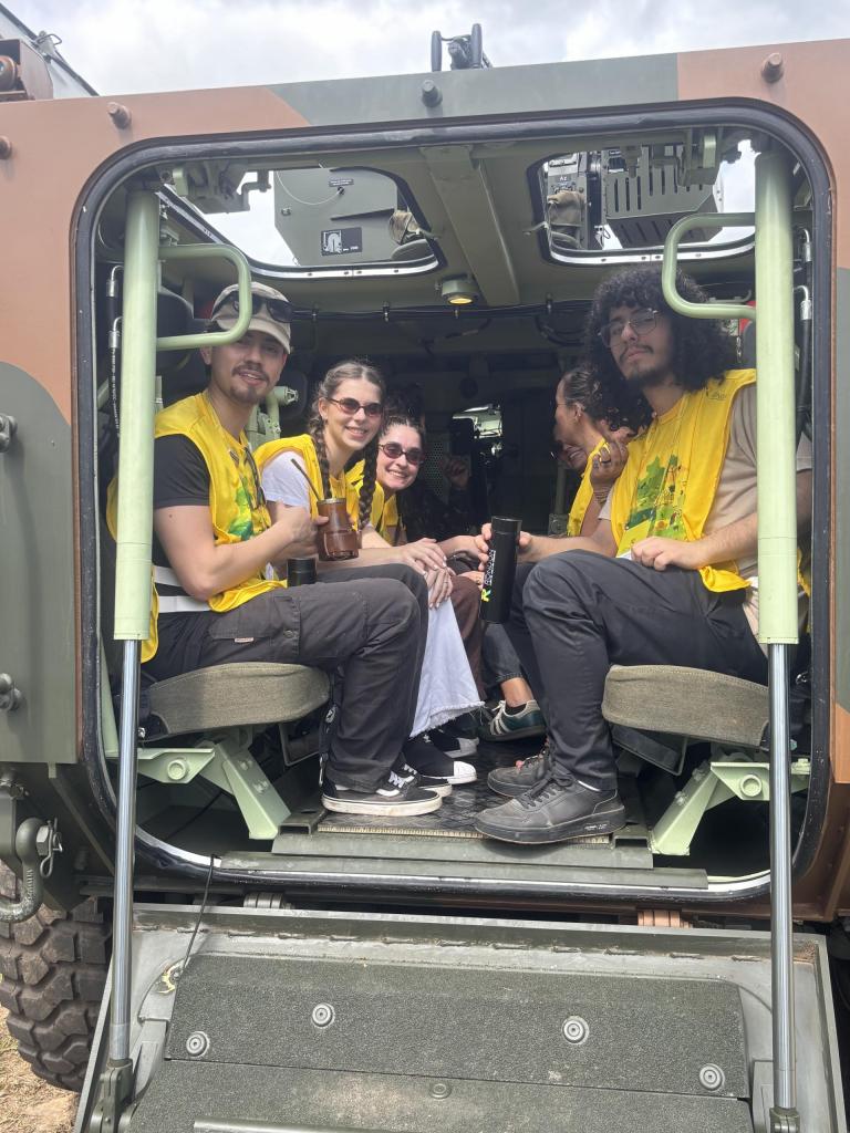 Fotografia colorida na vertical de estudantes que integram a equipe do Projeto Rondon da UFSM no interior de uma viatura militar durante deslocamento na centésima operação do Projeto Rondon. Seis estudantes vestem coletes amarelos de identificação e estão sentados em bancos laterais distribuídos nos dois lados do veículo. Cinco deles olham para a câmera e dois seguram recipientes para bebidas. A imagem é registrada a partir da abertura traseira da viatura do 52º Batalhão de Infantaria de Selva, permitindo visualizar a estrutura interna do veículo, as escotilhas superiores abertas e o céu ao fundo.
