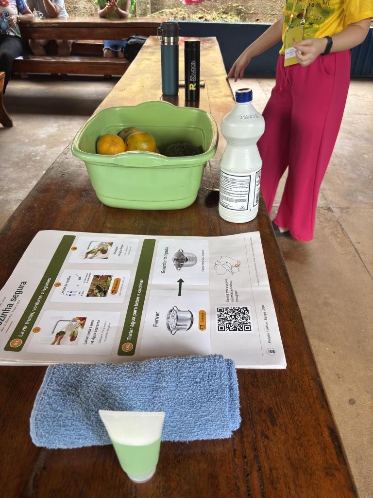 Fotografia colorida na vertical de uma oficina sobre higiene e segurança alimentar realizada por integrantes do Projeto Rondon da UFSM. Em primeiro plano, sobre uma mesa de madeira, há um material didático ilustrado, uma bacia plástica verde com frutas, um frasco branco, um pano azul e uma escova de limpeza. Ao fundo, uma integrante uniformizada do projeto aparece parcialmente ao lado da mesa enquanto conduz a atividade. Mais atrás, outras pessoas permanecem sentadas em bancos de madeira.