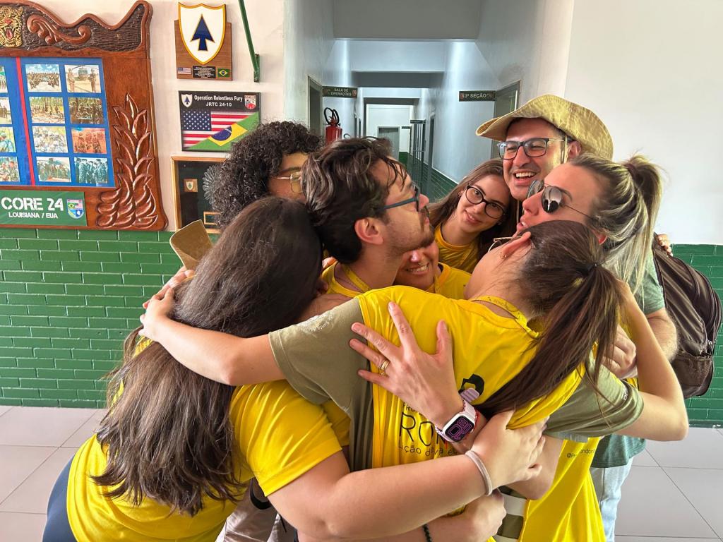 Foto colorida na horizontal. Em um corredor, oito estudantes e dois professores da equipe do Projeto Rondon pela UFSM aparecem reunidos em um abraço coletivo, em um momento de confraternização. O grupo está próximo à parede, que possui revestimento verde na parte inferior e fotografias, placas e símbolos relacionados a atividades militares e ao Projeto Rondon na parte superior. Os participantes vestem camisetas amarelas e verdes, e alguns usam óculos. Ao fundo, há portas e placas de identificação das salas. A cena transmite descontração, união e integração entre os integrantes da equipe.